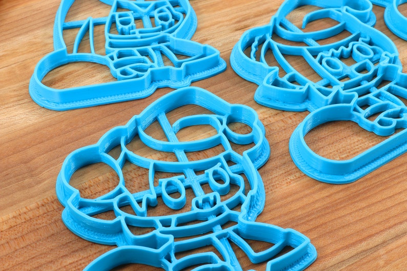 May include: Three blue plastic cookie cutters shaped like cartoon characters. The cutters are on a wooden surface.
