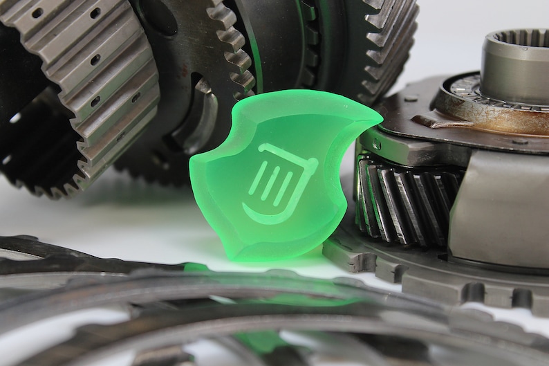 May include: A translucent green shield-shaped object with a white harp symbol, set against a backdrop of various metal gears and mechanical components. The gears are in shades of grey and silver.