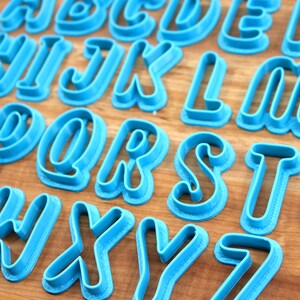 May include: A set of 26 blue plastic cookie cutters shaped like the letters of the alphabet. The letters are in a cursive font and have a raised edge.
