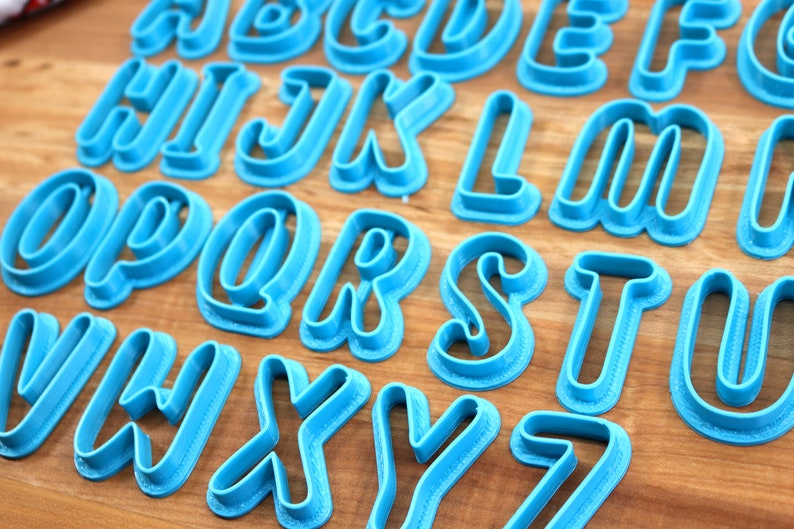 May include: A set of 26 blue plastic cookie cutters shaped like the letters of the alphabet. The letters are in a cursive font and have a raised edge.