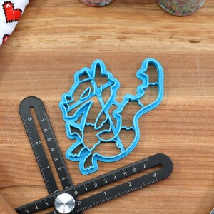 May include: A blue plastic cookie cutter shaped like a cartoon dragon. The cutter is on a wooden surface with a ruler and a rolling pin.