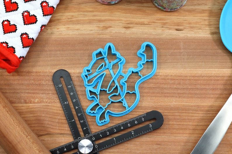 May include: A blue plastic cookie cutter shaped like a cartoon dragon. The cutter is on a wooden surface with a ruler and a rolling pin.