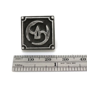 May include: A silver metal pin with a square design featuring a stylized symbol of three overlapping crescent shapes. The pin is approximately 2 cm wide.