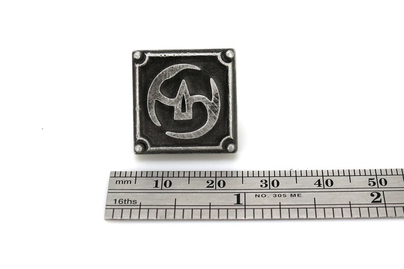 May include: A silver metal pin with a square design featuring a stylized symbol of three overlapping crescent shapes. The pin is approximately 2 cm wide.