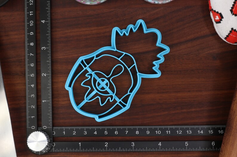 May include: A blue plastic cookie cutter shaped like a pineapple with a detailed design. The cutter is on a wooden surface with a ruler for scale.
