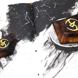 May include: Two brown resin geometric shapes with a gold symbol in the center. The shapes are on a page of a notebook with black ink drawings.
