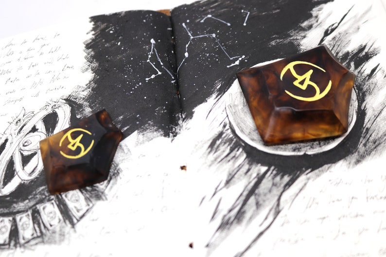 May include: Two brown resin geometric shapes with a gold symbol in the center. The shapes are on a page of a notebook with black ink drawings.