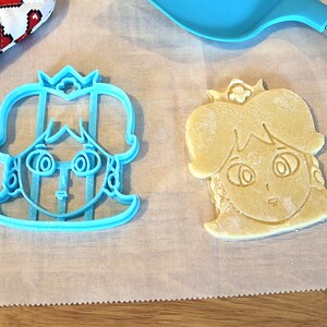 May include: A blue plastic cookie cutter shaped like a princess with a crown, next to a light brown cookie that has been cut out with the cutter.