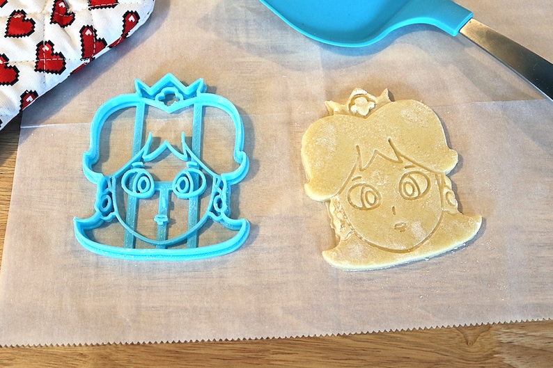 May include: A blue plastic cookie cutter shaped like a princess with a crown, next to a light brown cookie that has been cut out with the cutter.