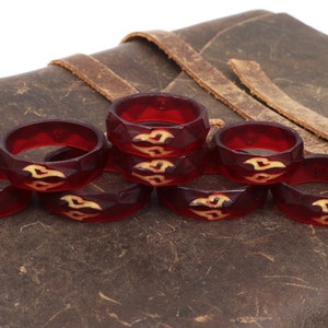 May include: Eight red resin rings with a geometric design and a cream-colored pattern. The rings are stacked on a brown leather surface.