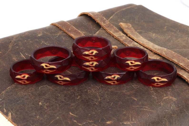 May include: Eight red resin rings with a geometric design and a cream-colored pattern. The rings are stacked on a brown leather surface.