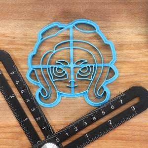 May include: A blue plastic cookie cutter shaped like a cartoon character with large eyes and hair. The cutter is on a wooden surface.