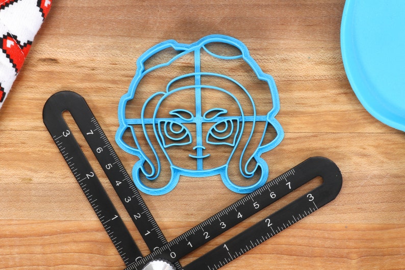 May include: A blue plastic cookie cutter shaped like a cartoon character with large eyes and hair. The cutter is on a wooden surface.