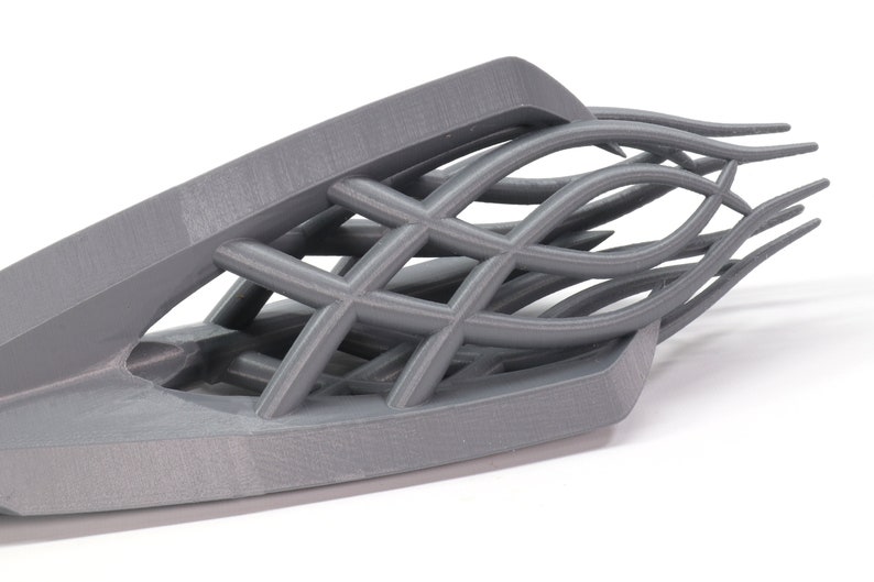 May include: A gray 3D printed object with a geometric design. The object has a curved shape and a series of intersecting lines that create a lattice-like pattern.