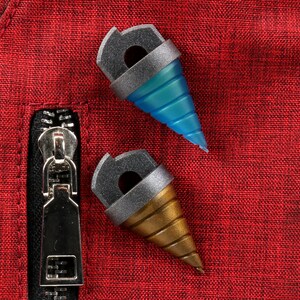 May include: Two drill-shaped accessories with gray and gold accents, one blue and one gold, rest on a red textured surface. The accessories have a cylindrical top with a hole and a conical drill bit. A silver zipper is visible.