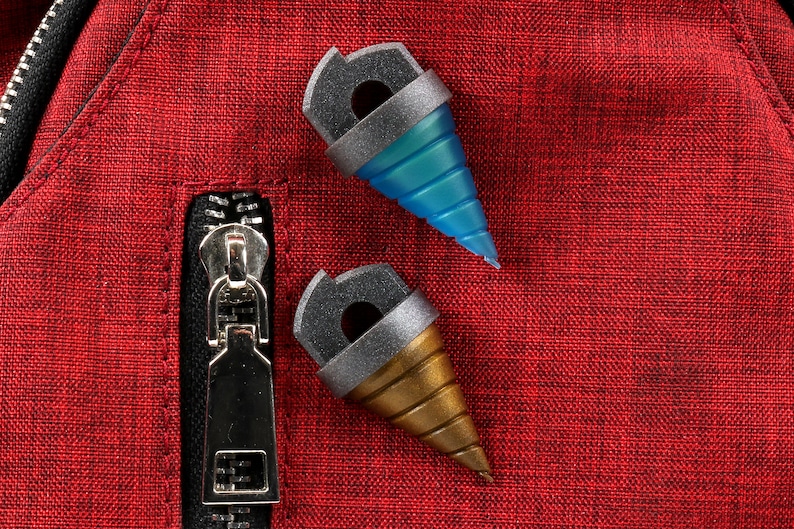 May include: Two drill-shaped accessories with gray and gold accents, one blue and one gold, rest on a red textured surface. The accessories have a cylindrical top with a hole and a conical drill bit. A silver zipper is visible.
