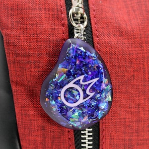 Puede incluir: Un llavero de resina azul y morado con un dise&ntilde;o de llama blanca. El llavero est&aacute; unido a un tirador de cremallera rojo.