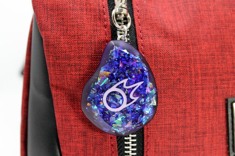 Puede incluir: Un llavero de resina azul y morado con un dise&ntilde;o de llama blanca. El llavero est&aacute; unido a un tirador de cremallera rojo.