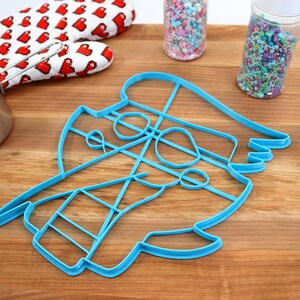 May include: A blue plastic cookie cutter shaped like a cartoon pirate with a hat, eye patch, and beard. The cutter is on a wooden surface with sprinkles in jars and a red oven mitt.