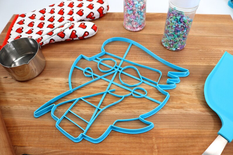 May include: A blue plastic cookie cutter shaped like a cartoon pirate with a hat, eye patch, and beard. The cutter is on a wooden surface with sprinkles in jars and a red oven mitt.