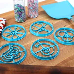May include: Six blue plastic cookie cutters with different designs on a wooden cutting board. There are two jars of sprinkles in the background.