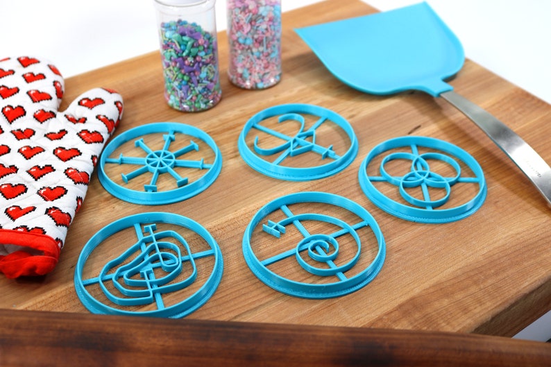 May include: Six blue plastic cookie cutters with different designs on a wooden cutting board. There are two jars of sprinkles in the background.