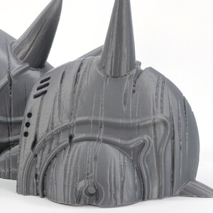 May include: A 3D printed gray helmet with a textured surface and a pointed horn on top. The helmet is made in multiple pieces and has a design that resembles a warrior's helmet.