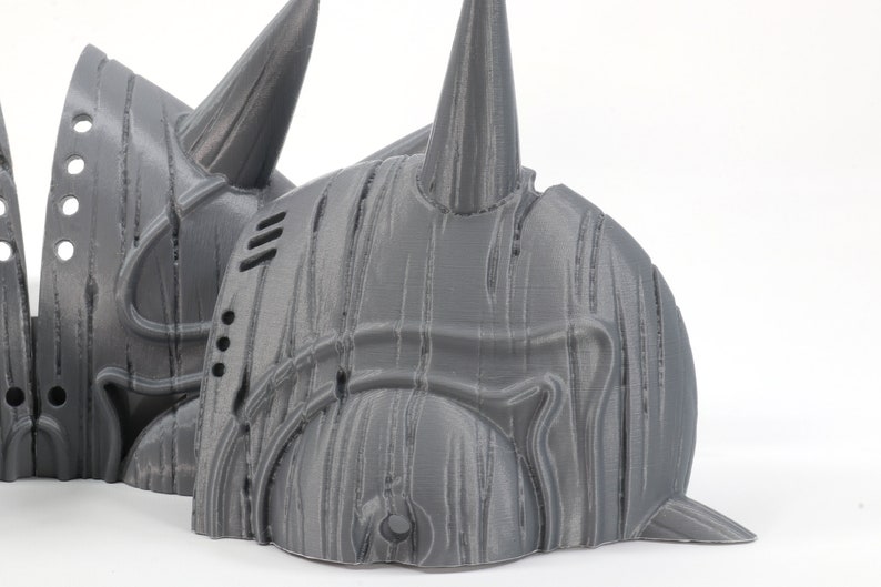May include: A 3D printed gray helmet with a textured surface and a pointed horn on top. The helmet is made in multiple pieces and has a design that resembles a warrior's helmet.