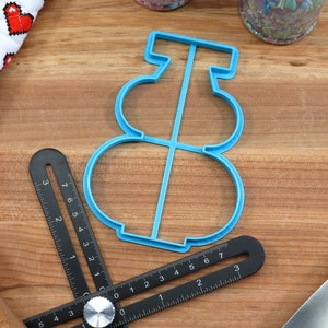 May include: A blue plastic cookie cutter shaped like a snowman. The cutter has three circles for the snowman's body and a small rectangle for the hat.