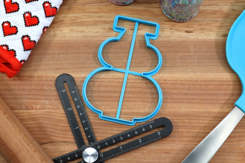 May include: A blue plastic cookie cutter shaped like a snowman. The cutter has three circles for the snowman's body and a small rectangle for the hat.