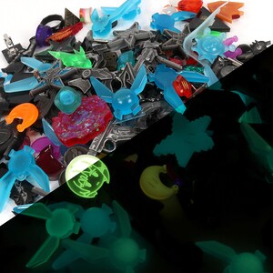 May include: A collection of glow-in-the-dark charms in various shapes and colors, including blue, green, and yellow. The charms are arranged on a white surface and a black surface to show how they glow in the dark.