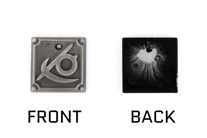 May include: A silver metal charm with a stylized eye and a curved blade design on the front. The back of the charm is black with a silver-colored design.