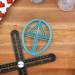 May include: A blue plastic cookie cutter shaped like a triangle inside a circle. The cutter is on a wooden surface with a black ruler.
