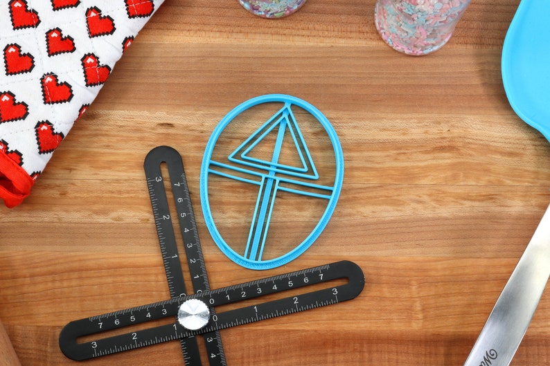 May include: A blue plastic cookie cutter shaped like a triangle inside a circle. The cutter is on a wooden surface with a black ruler.