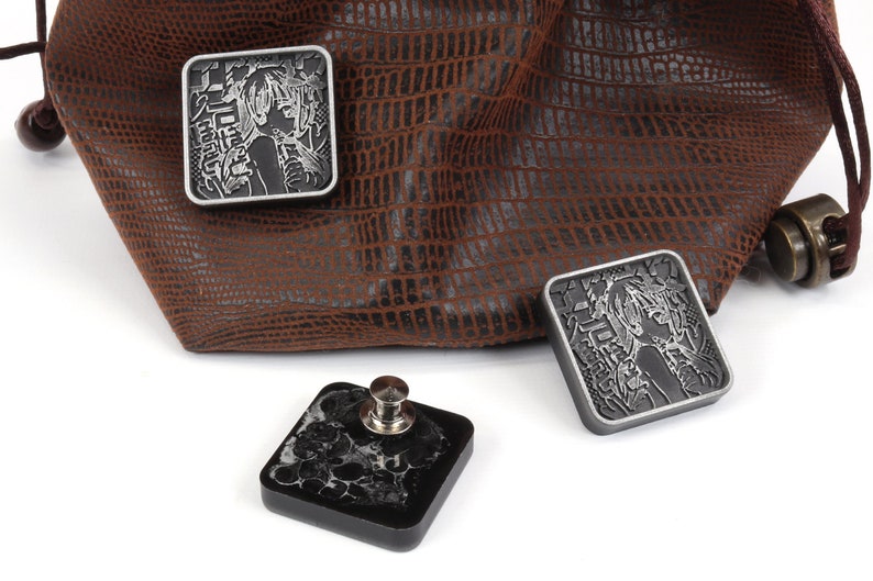 Puede incluir: Tres dados cuadrados de metal con acabado plateado y un dise&ntilde;o grabado de una mujer con texto japon&eacute;s. Los dados est&aacute;n sobre una superficie blanca junto a una bolsa de cuero marr&oacute;n.
