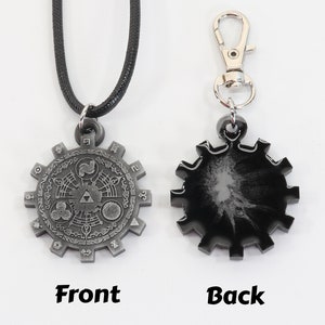 May include: A silver-colored pendant necklace with a detailed design of a circular gear with symbols and numbers. The back of the pendant is black with a silver-colored design. The necklace is on a black cord.