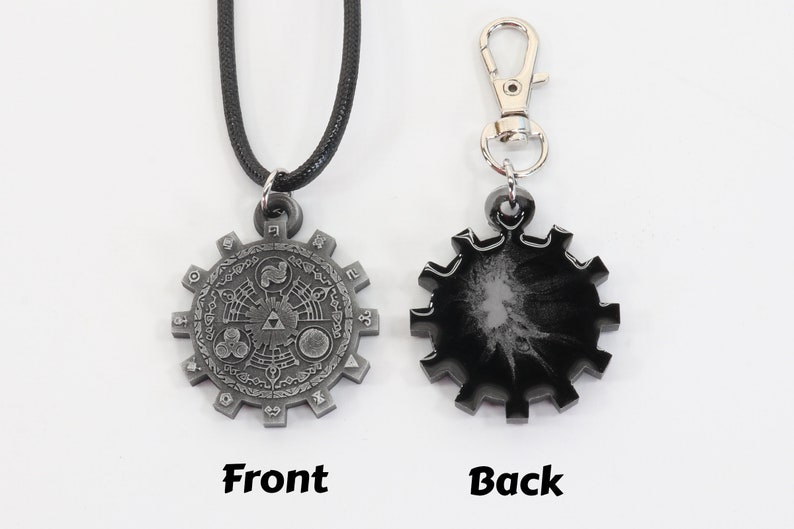 May include: A silver-colored pendant necklace with a detailed design of a circular gear with symbols and numbers. The back of the pendant is black with a silver-colored design. The necklace is on a black cord.