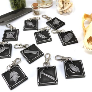 May include: Ten silver keychains with different symbols on each. The symbols include a fish, a feather, a cloud, a crown, a leaf, a hammer, a helmet, a knife, a crescent moon, and a wrench. The keychains are on a white surface.