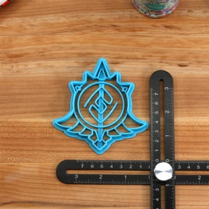 May include: A blue plastic cookie cutter shaped like a stylized symbol with a sword and a circle. The cutter is about 2 inches in diameter and is sitting on a wooden surface.