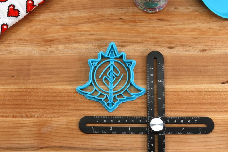 May include: A blue plastic cookie cutter shaped like a stylized symbol with a sword and a circle. The cutter is about 2 inches in diameter and is sitting on a wooden surface.