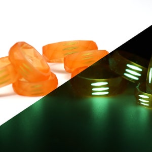May include: A group of orange glow-in-the-dark rings with green stripes. The rings are stacked on top of each other on a white surface. The rings are glowing green in the dark.