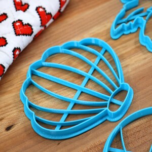 May include: Three blue plastic cookie cutters shaped like a seashell, a palm tree, and a hot air balloon. The cutters are on a wooden surface.