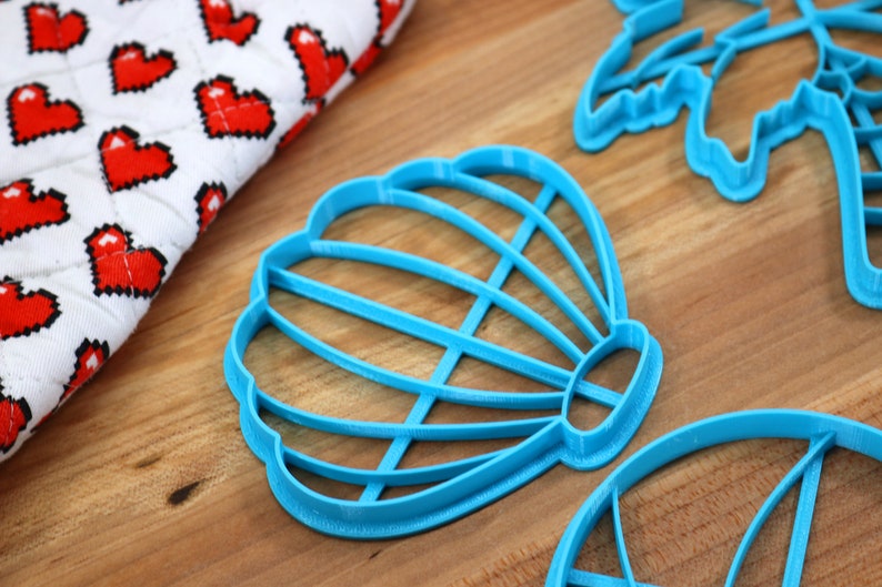 May include: Three blue plastic cookie cutters shaped like a seashell, a palm tree, and a hot air balloon. The cutters are on a wooden surface.