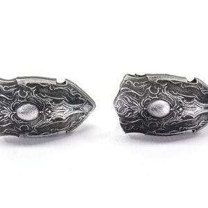 May include: Two silver-coloured shield-shaped pendants with intricate designs. The pendants have a raised, circular centre and are detailed with a swirling pattern.