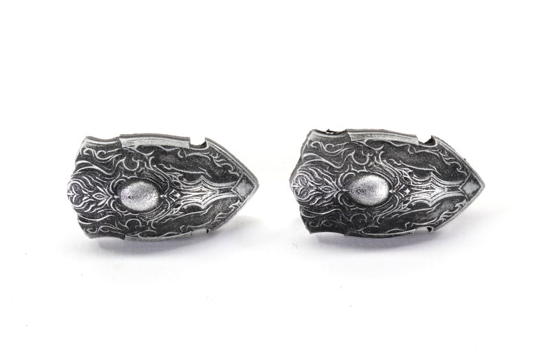 May include: Two silver-coloured shield-shaped pendants with intricate designs. The pendants have a raised, circular centre and are detailed with a swirling pattern.