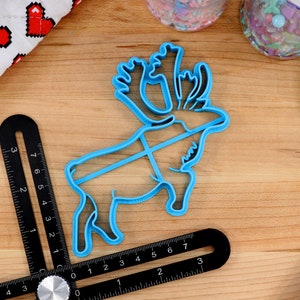 May include: A blue plastic reindeer cookie cutter with intricate details on the antlers. The cutter is on a wooden surface with a ruler and other baking supplies.
