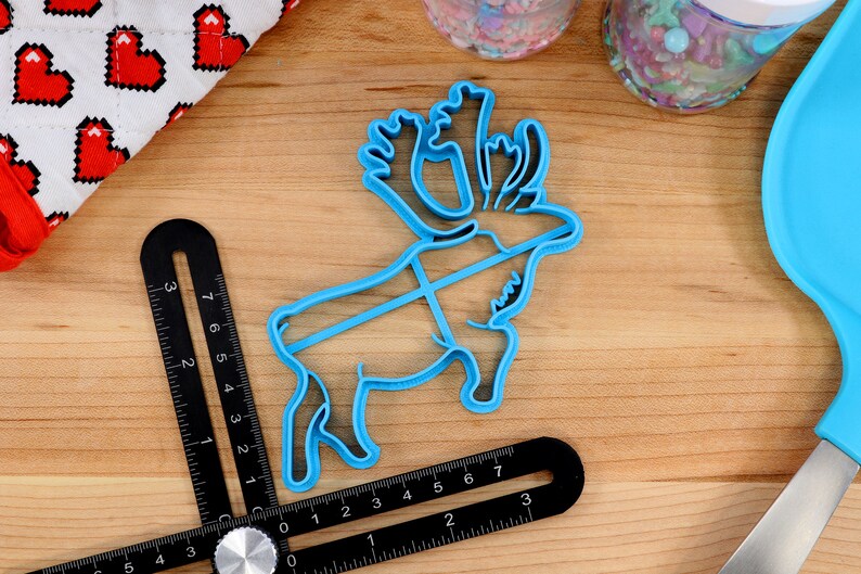May include: A blue plastic reindeer cookie cutter with intricate details on the antlers. The cutter is on a wooden surface with a ruler and other baking supplies.