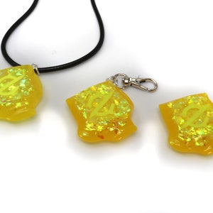 May include: Three yellow resin charms with a glittery finish. The charms feature a stylized letter "Z" design. One charm is on a black cord necklace, another is on a silver key chain, and the third charm is on a white surface.