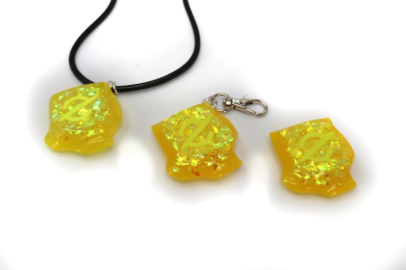 May include: Three yellow resin charms with a glittery finish. The charms feature a stylized letter "Z" design. One charm is on a black cord necklace, another is on a silver key chain, and the third charm is on a white surface.