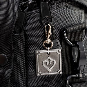 May include: Black zippered bag with a silver metal charm featuring a stylized heart design. The bag has a black strap with a metal buckle.